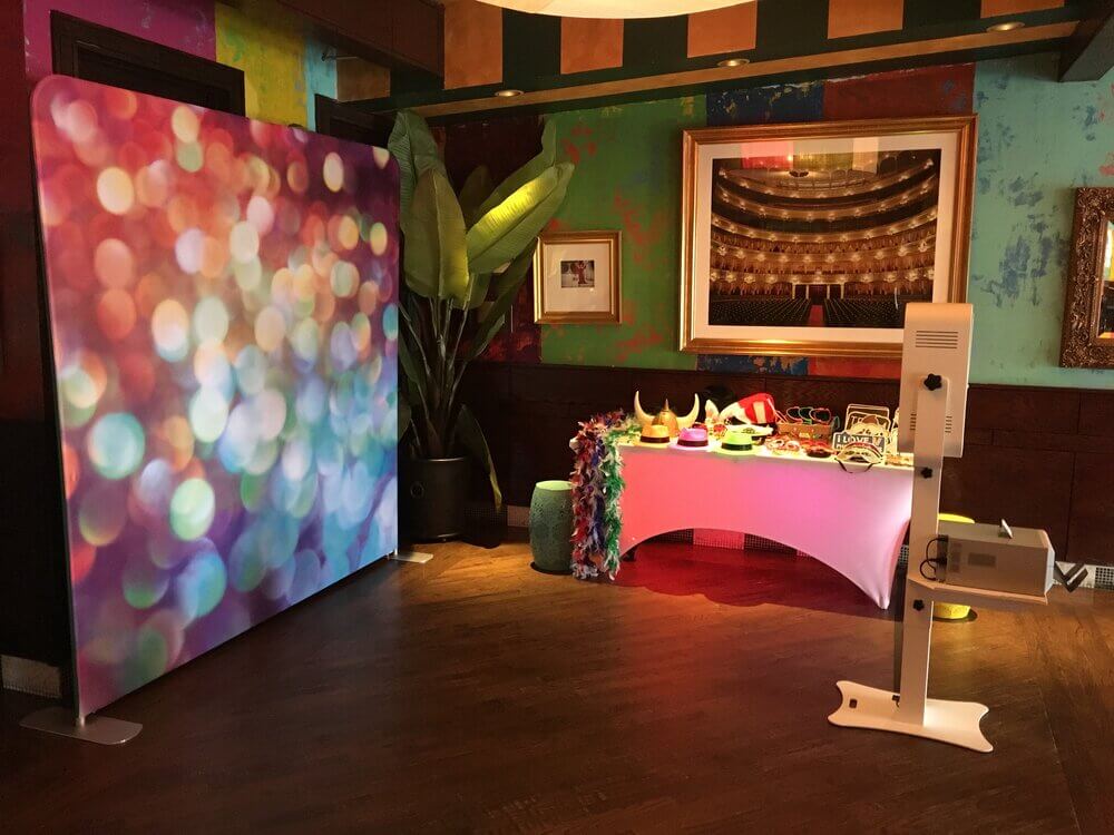 A colorful event setup with a bokeh-patterned partition, a table with hats and masks, and a digital kiosk.