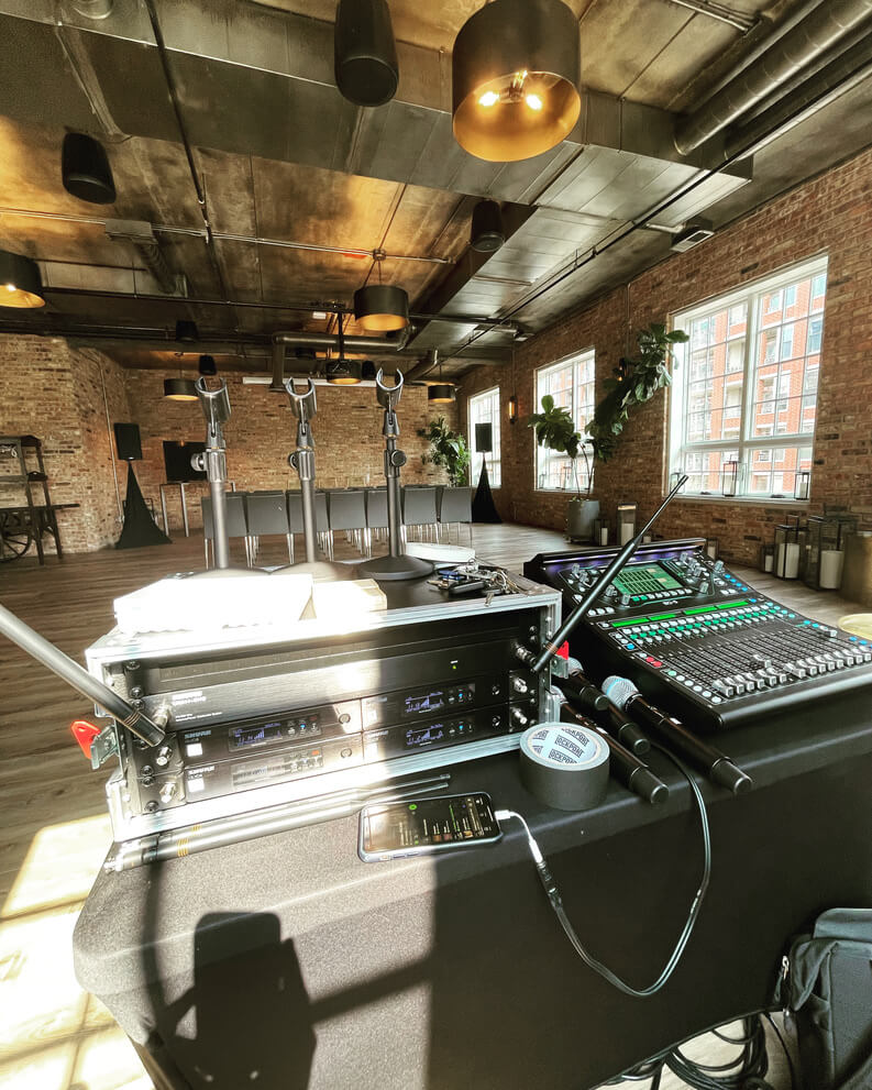 A dj setup with mixers and turntables in an industrial-style venue with exposed brick walls and large windows.