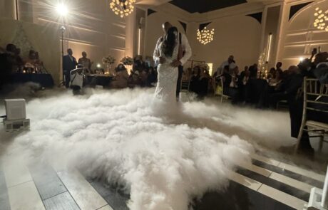 Couple sharing a first dance on a dance floor surrounded by a cloud effect with guests watching in the background.