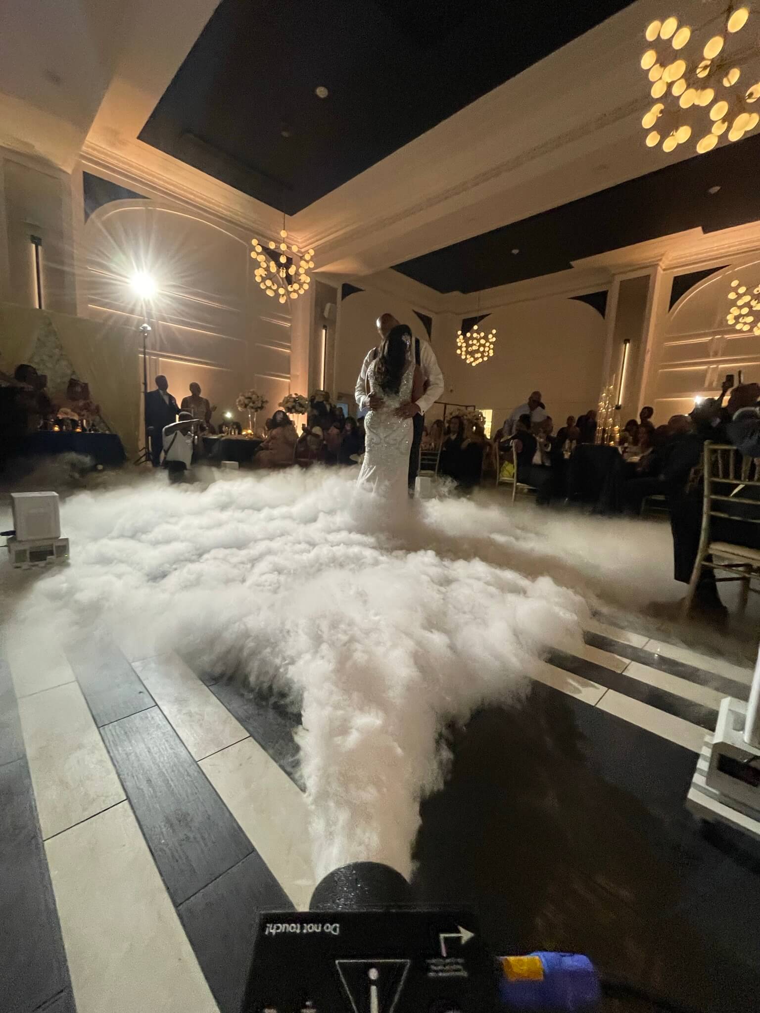 Couple sharing a first dance on a dance floor surrounded by a cloud effect with guests watching in the background.