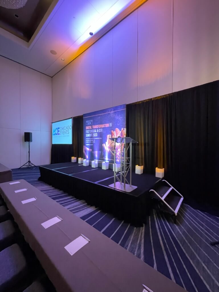 A speaker presenting at a conference in a room with stage lighting and a projector screen displaying text.