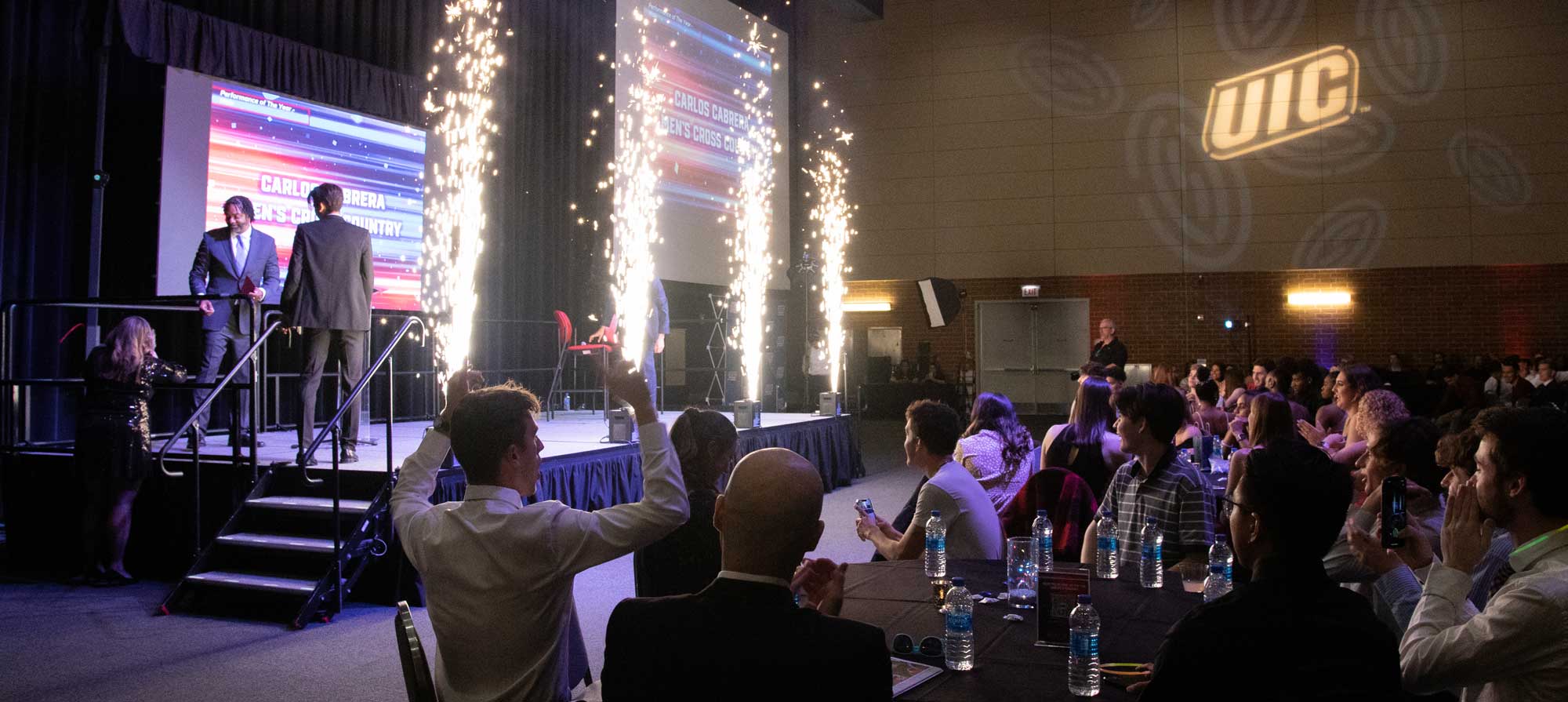 Two individuals on a stage with pyrotechnic effects as an audience watches at an indoor event.