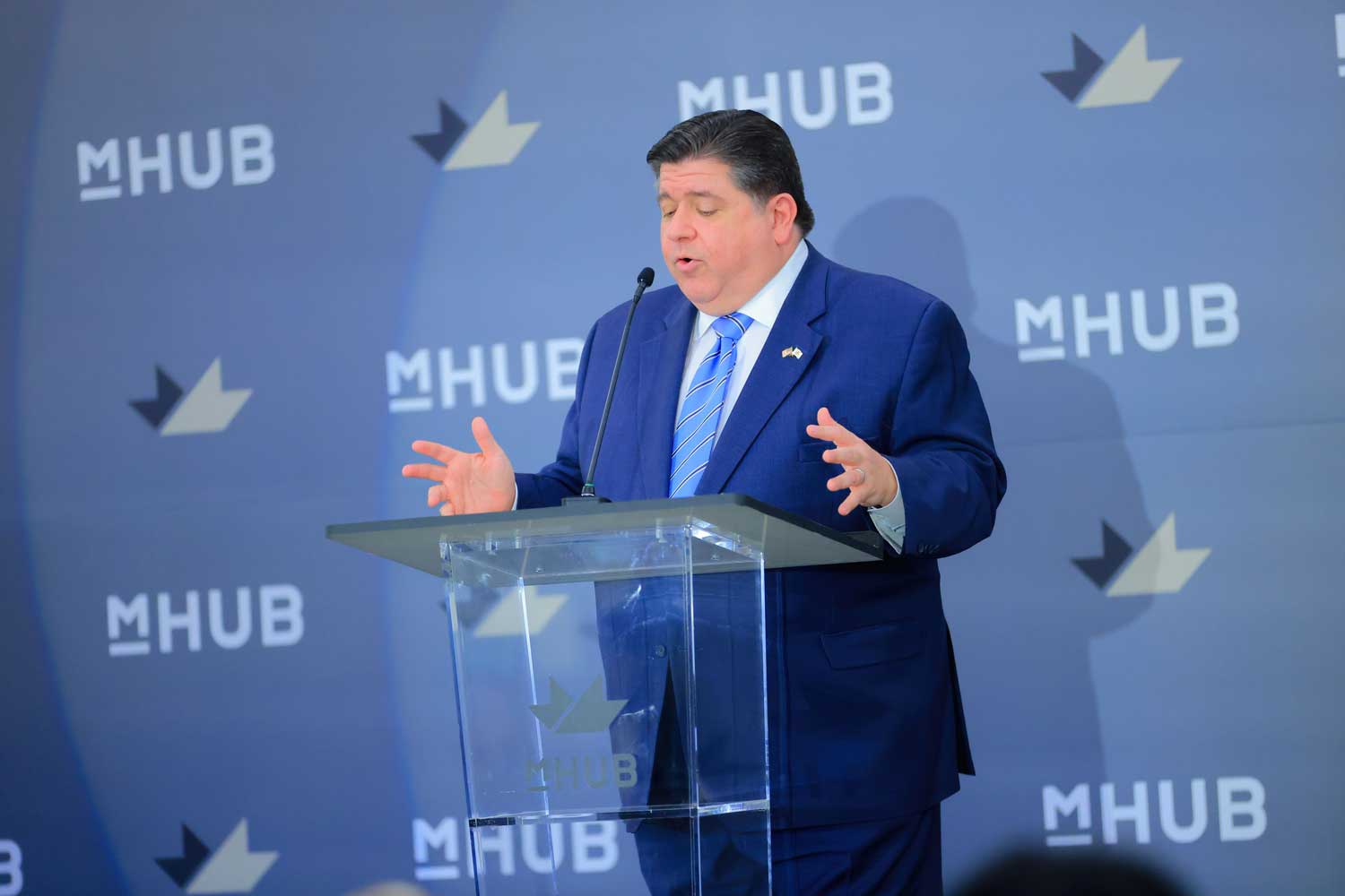 A man in a suit speaking at a podium with the "mhub" logo in the background.