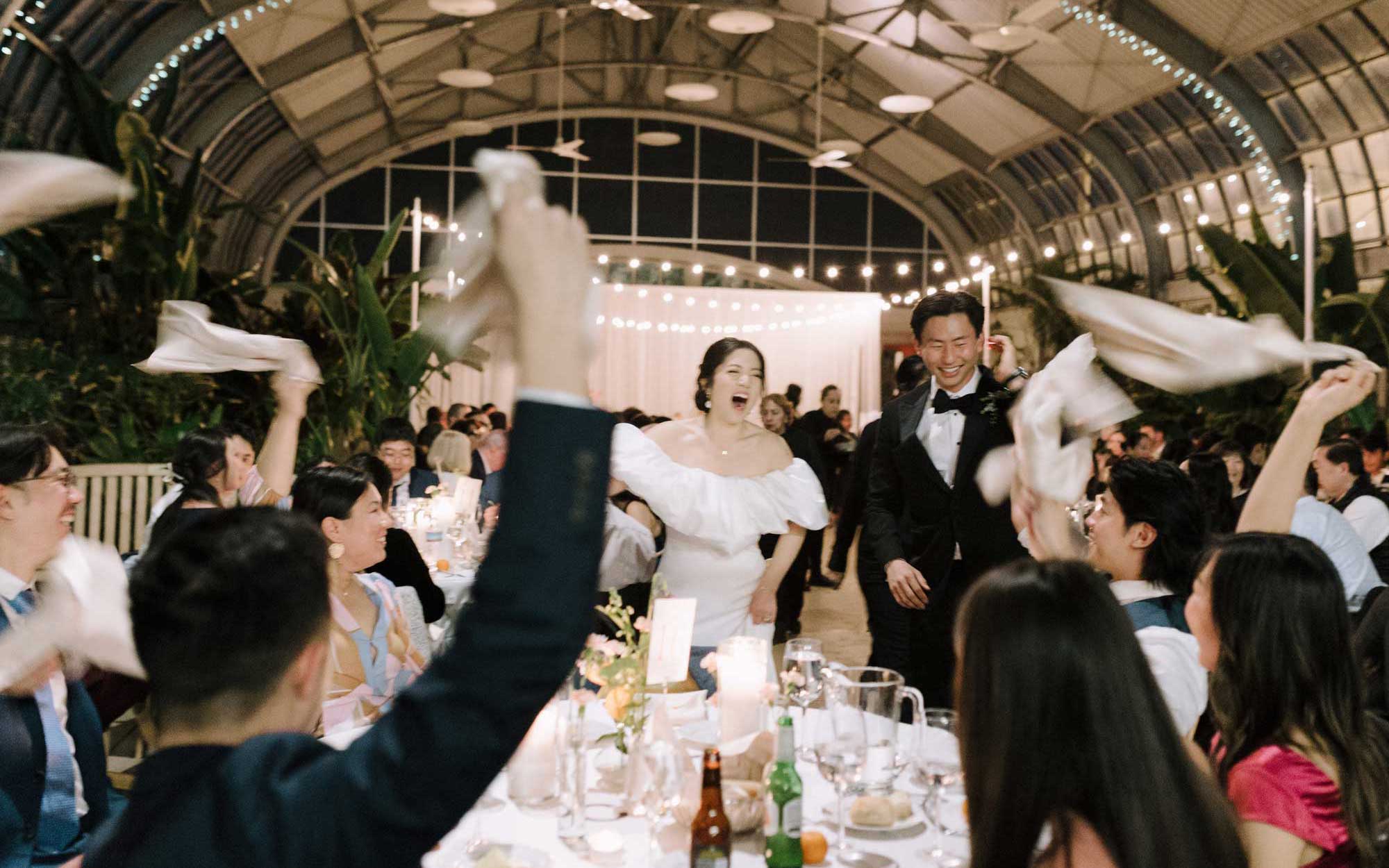 A happy couple walks through a cheering crowd at a wedding reception while guests wave their napkins in celebration.
