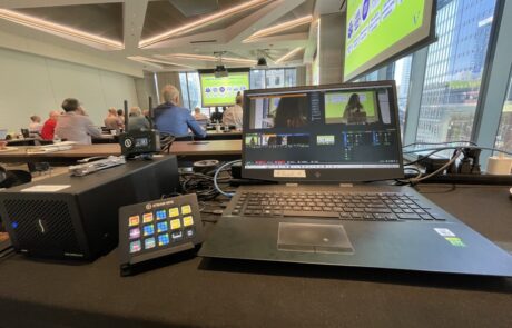 A control station with a laptop overseeing a conference room with attendees facing a presentation screen.
