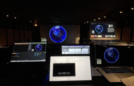 A laptop on a desk with multiple monitor screens displaying earth graphics in a dimly lit control room.