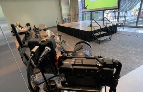 A video camera set up in a conference room with presentation screens in the background.