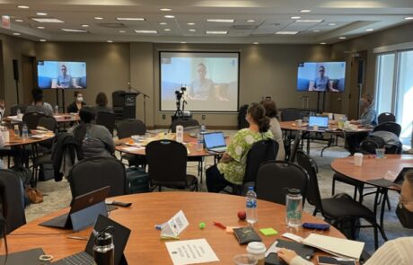 A group of professionals attending a conference in a meeting room with a speaker presenting on a projector screen.