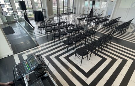 An event space with chequered flooring prepared with rows of chairs and audio equipment in the foreground.