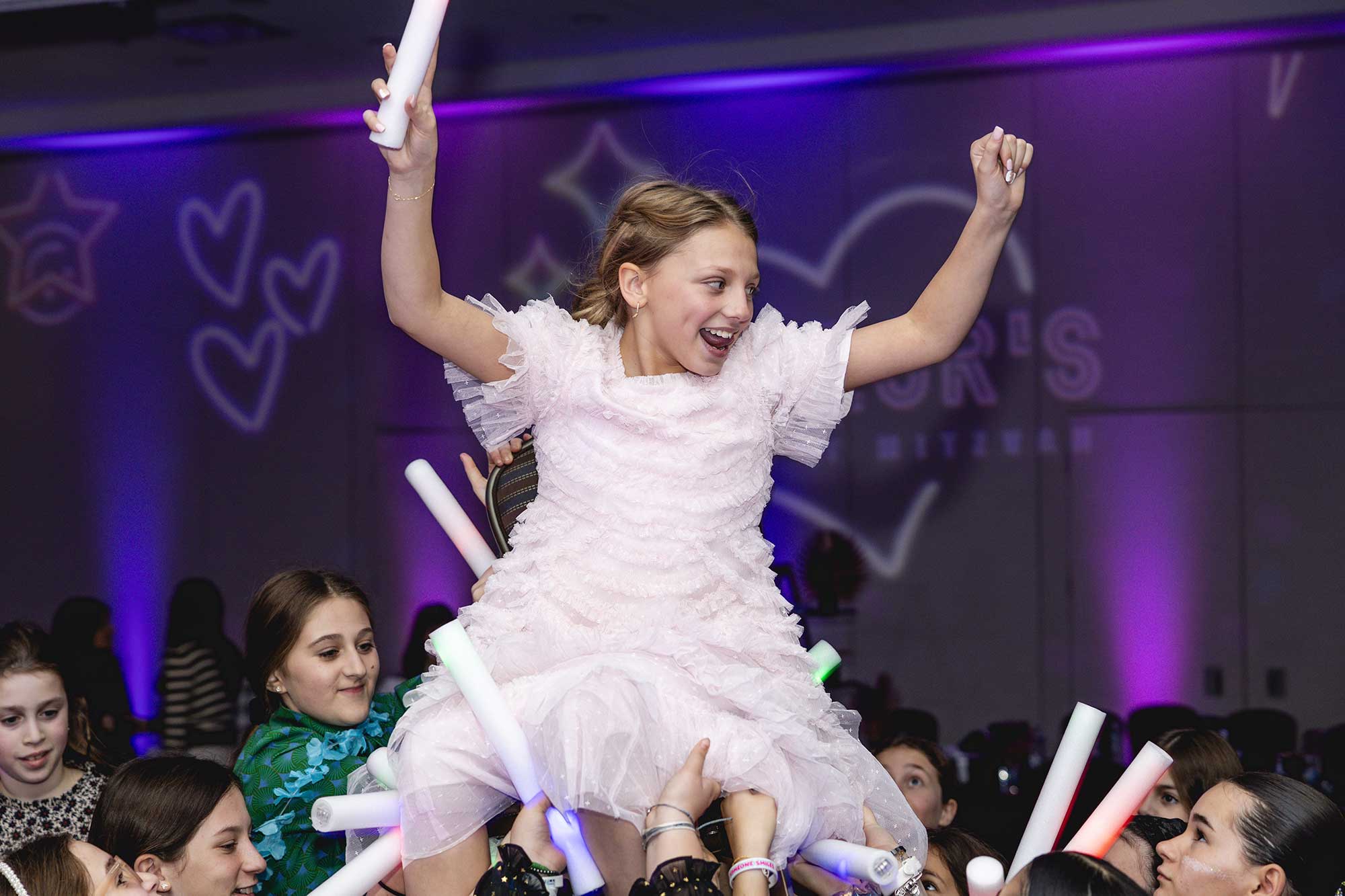 A joyful girl being lifted in the air on a chair amidst a celebratory event with other children surrounding her with glowing sticks.