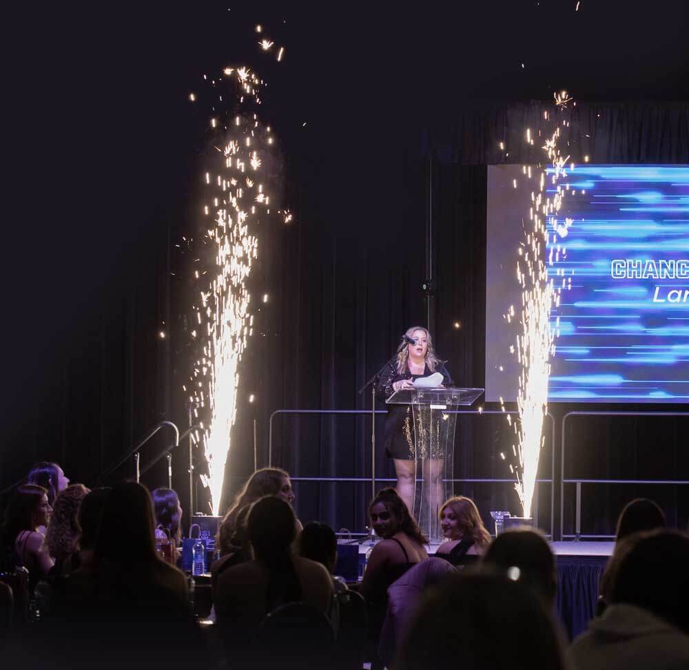 Woman delivering a speech at a podium with pyrotechnic effects on stage.