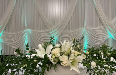 Floral centerpiece with white flowers and greenery on a table against a draped fabric background with ambient lighting.