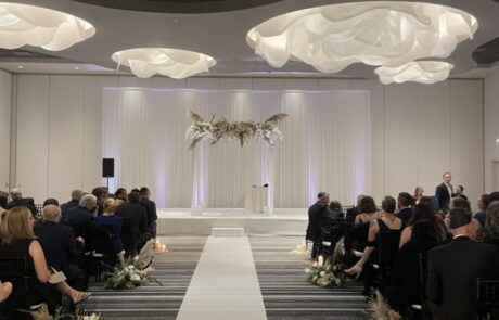 An indoor wedding ceremony with guests seated on either side of a white aisle leading to an altar adorned with floral decorations.