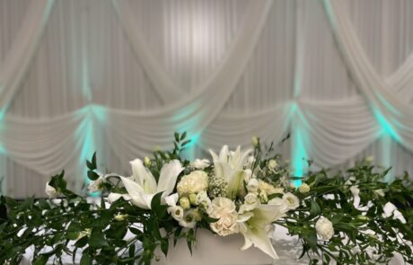 A floral centerpiece arrangement with white flowers and greenery on a table, with a draped white curtain and blue uplighting in the background.