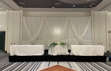 A draped white backdrop with a floral centerpiece on a skirted table at an event hall.