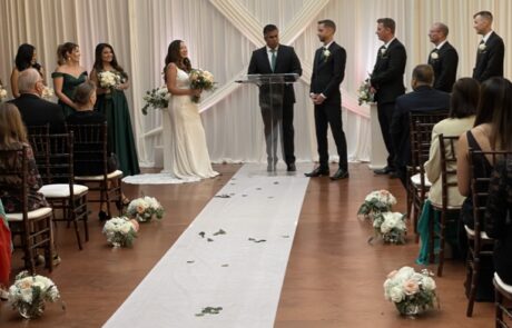 A wedding ceremony in progress inside a venue with wooden beams, a bride and groom standing at the altar, and guests seated on either side of a white aisle runner.