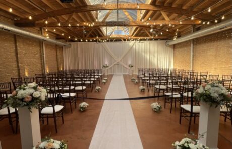 An elegantly decorated wedding aisle with a white carpet, chairs lined up on either side, and floral arrangements in a venue with wooden beams and brick walls.