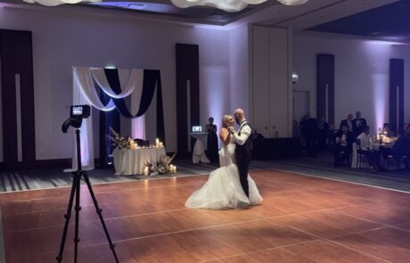 A couple sharing a dance in a banquet hall with guests looking on and a camera set up in the foreground.