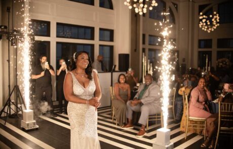 A joyful woman in an elegant dress stands amidst sparkling pyrotechnics at an indoor event, with smiling guests looking on.