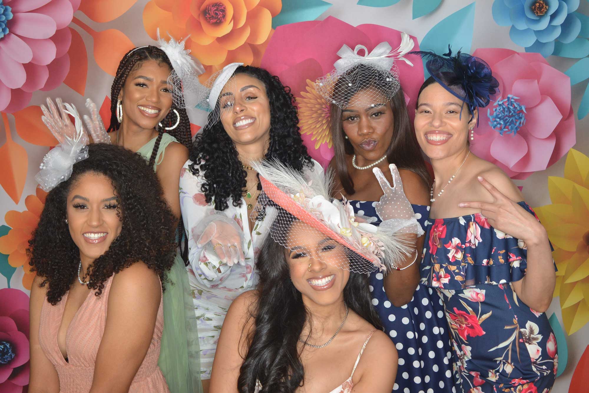 A group of joyful women dressed in festive attire with floral hats posing together in front of a colorful flower backdrop.