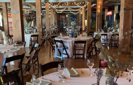 A decorated event space with tables set for a celebration, featuring string lights and wooden beams.