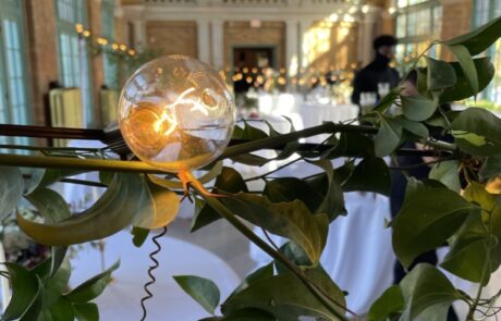 Elegant event setting with a focus on a table decoration featuring a clear orb and greenery, with a chandelier-lit hall in the background.