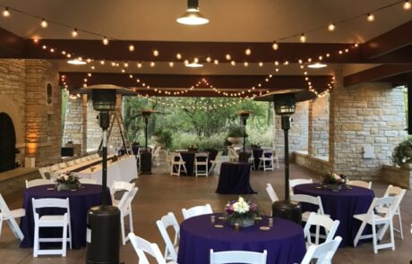 An outdoor event space with string lights, white chairs, and purple tablecloths set up for a gathering.
