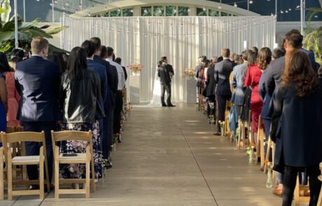 Guests standing at a wedding ceremony inside a greenhouse with the photographer capturing the moment.