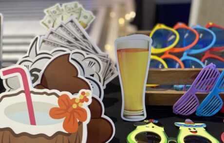Assorted party props including glasses with fun designs, a paper cutout of a beer mug, and tropical-themed decorations on a table with a blurred event venue in the background.