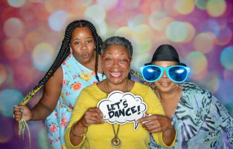 Three women posing with party props against a colorful backdrop, with the central figure holding a "let's dance!" sign.