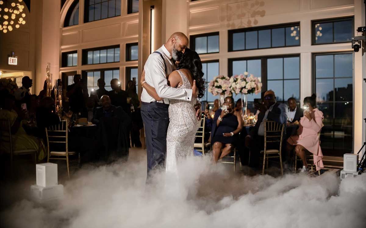 A couple shares a dance in a romantic setting with a fog effect on the dance floor, surrounded by seated guests.