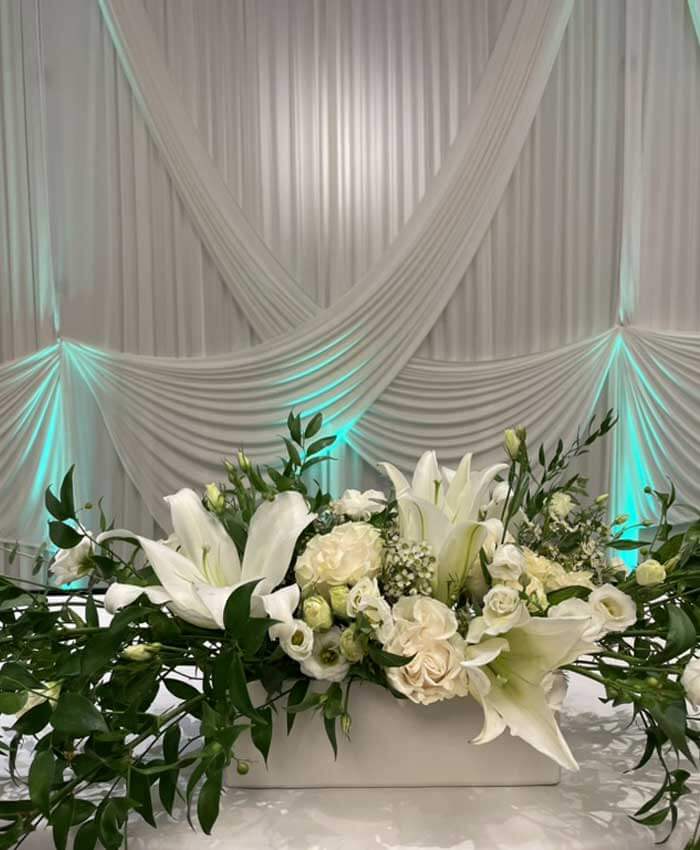Floral arrangements with white flowers on a table in front of a draped backdrop with teal accent lighting.