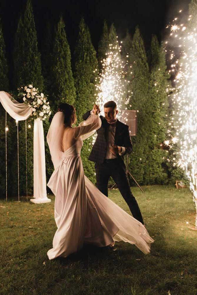 A bride and groom dance outdoors at night with sparklers in the background, creating a festive atmosphere.