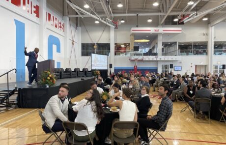 Speaker addressing an audience at an indoor event with attendees seated at round tables.