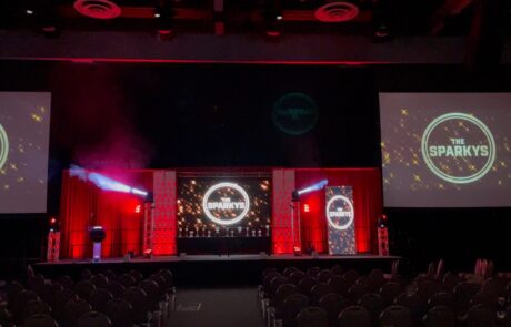 Stage setup for 'the sparkys' event with empty seats, lighting, and two large projection screens displaying the event's logo.