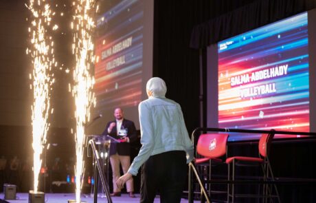 A person approaching a stage with pyrotechnics as their name is displayed on the screen, possibly at an award ceremony or event.