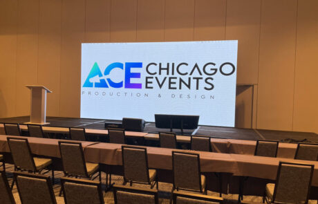 A conference room setup with chairs facing a stage and a large screen displaying the "ace chicago events production & design" logo.