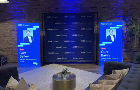 Event stage setup with two large screens displaying a presenter's name and photo, surrounded by promotional banners and casual seating in a room with brick walls and ambient lighting.